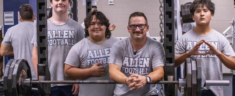 Four individuals wearing gray “Allen Football” T-shirts pose together inside a weight room. Three students stand behind a heavily loaded barbell on a squat rack, while an adult leans forward in front of the bar with his hands clasped. Weight plates, chains, mirrors, and additional gym equipment fill the background.