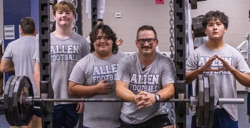 Four individuals wearing gray “Allen Football” T-shirts pose together inside a weight room. Three students stand behind a heavily loaded barbell on a squat rack, while an adult leans forward in front of the bar with his hands clasped. Weight plates, chains, mirrors, and additional gym equipment fill the background.