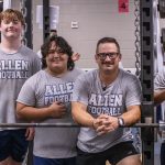 Four individuals wearing gray “Allen Football” T-shirts pose together inside a weight room. Three students stand behind a heavily loaded barbell on a squat rack, while an adult leans forward in front of the bar with his hands clasped. Weight plates, chains, mirrors, and additional gym equipment fill the background.
