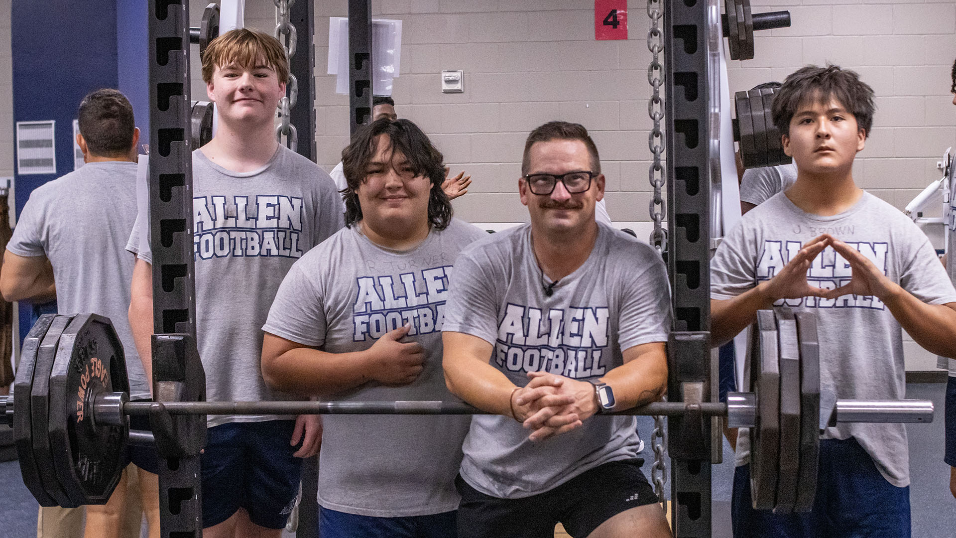 Four individuals wearing gray “Allen Football” T-shirts pose together inside a weight room. Three students stand behind a heavily loaded barbell on a squat rack, while an adult leans forward in front of the bar with his hands clasped. Weight plates, chains, mirrors, and additional gym equipment fill the background.
