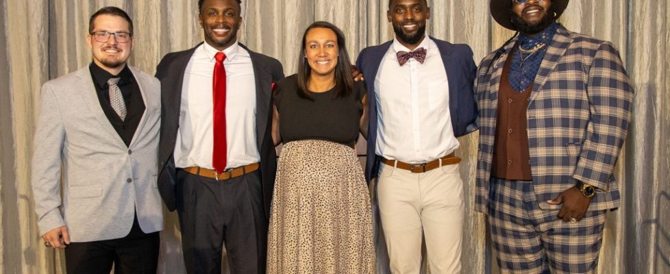 Five adults dressed in formal and semi-formal attire stand together smiling in front of a gray curtain backdrop.