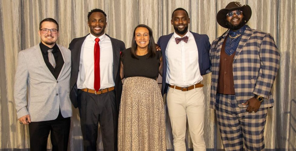 Five adults dressed in formal and semi-formal attire stand together smiling in front of a gray curtain backdrop.