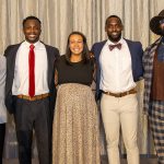 Five adults dressed in formal and semi-formal attire stand together smiling in front of a gray curtain backdrop.