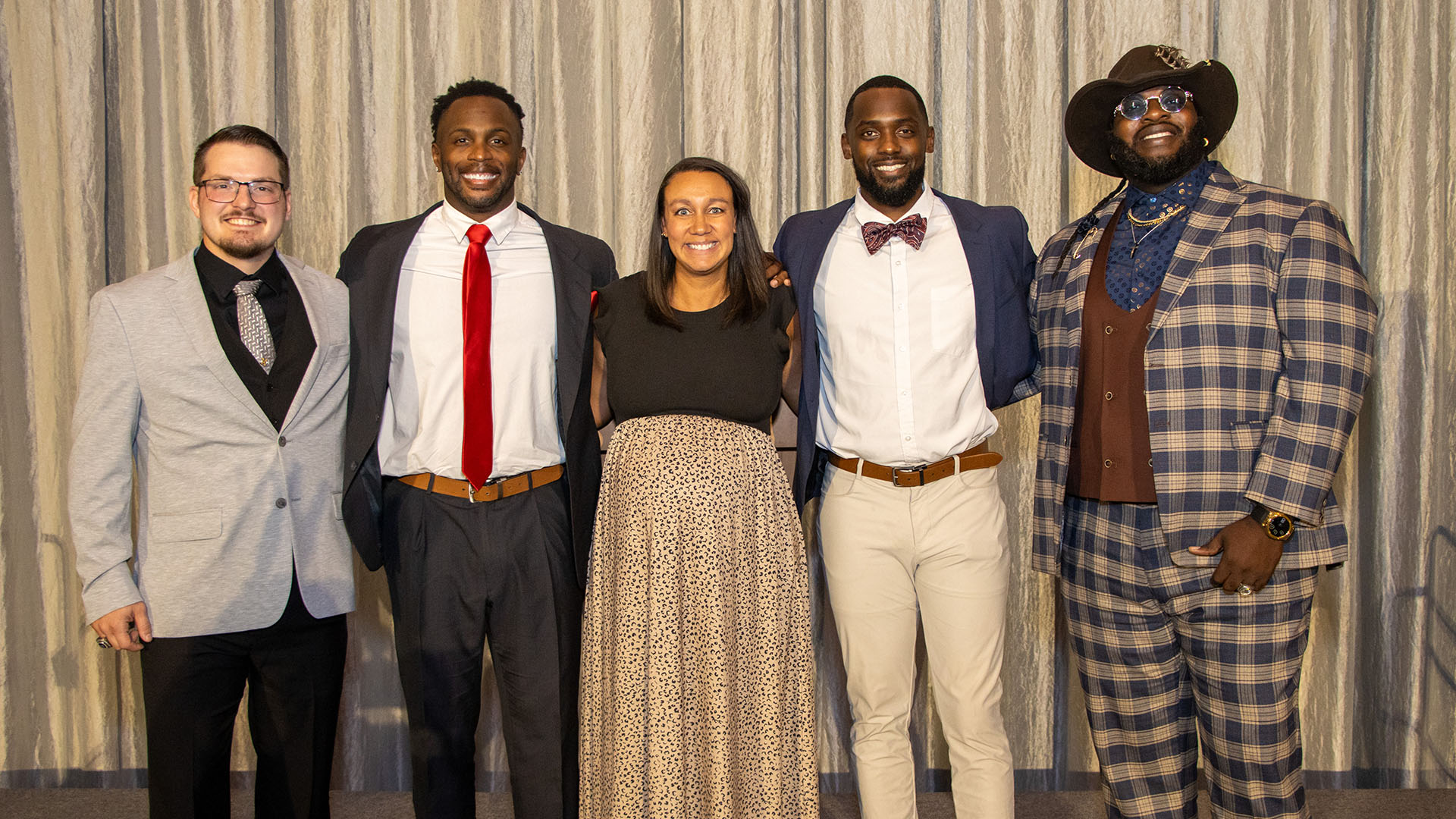 Five adults dressed in formal and semi-formal attire stand together smiling in front of a gray curtain backdrop.