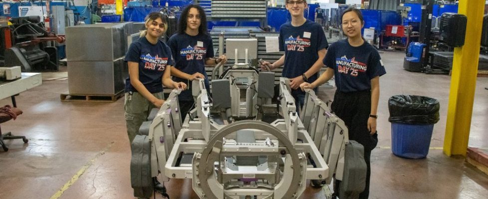 Four students wearing “Manufacturing Day ’25” shirts stand around a metal assembly fixture inside an industrial manufacturing facility.