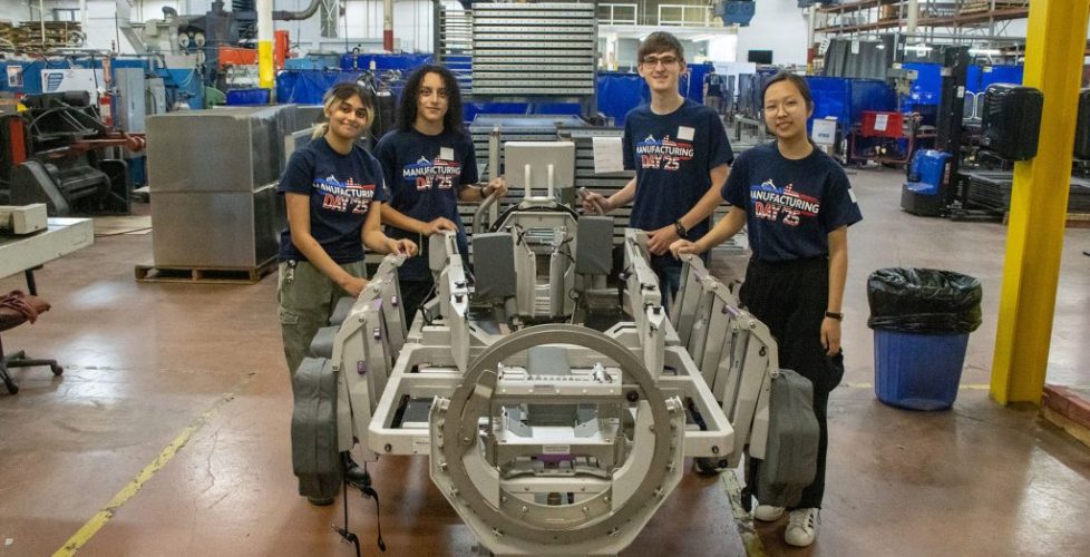 Four students wearing “Manufacturing Day ’25” shirts stand around a metal assembly fixture inside an industrial manufacturing facility.
