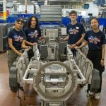 Four students wearing “Manufacturing Day ’25” shirts stand around a metal assembly fixture inside an industrial manufacturing facility.